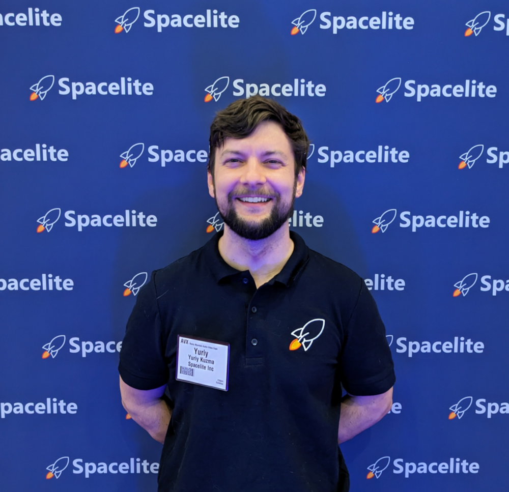 Picture of Yuriy Kuzma smiling, in front of a fabric sign with the Spacelite logo tiled across. Yuriy is wearing a navy blue polo with a small Spacelite rocket graphic embroidered over the heart.