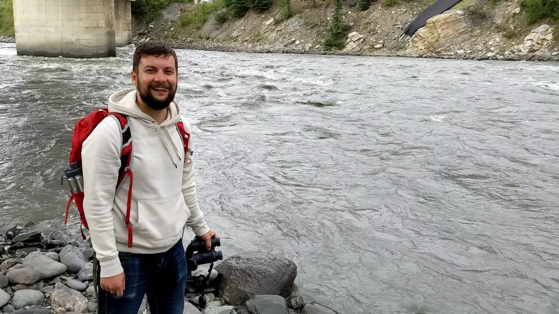 Picture of Yuriy Kuzma smiling, handing out by a river under a bridge, and holding binoculars. Yuriy is wearing a red backpack, white sweatshirt, and jeans.